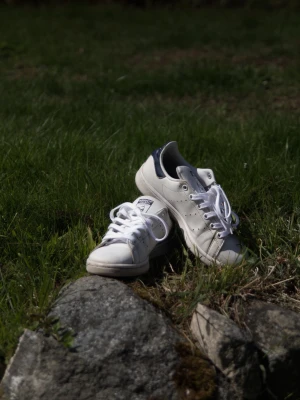 Adidas Stan Smith vita sneakers - Klassiska vita Adidas Stan Smith sneakers i läder med mörkblå detaljer på hälen. Skorna har vit sula, vita skosnören och en ikonisk logga på plösen. Perfekta för en clean och tidlös stil. Sparsamt använda och nya snören!        Boxen är tyvärr borta.                                     Priset kan sänkas vid snabb affär. 