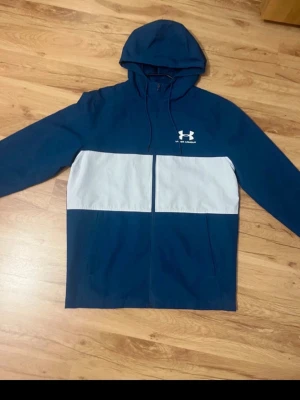 vindjacka från Under Armour - Snygg vindjacka från Under Armour i blått och vitt med huva och dragkedja framtill. Jackan har två sidofickor och en sportig look med logga på bröstet. Pris kan sänkas vid snabb affär. 
