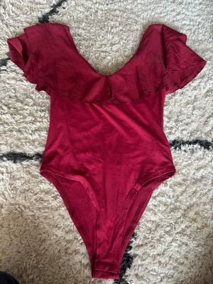 Vinröd off-shoulder body SHEIN - Snygg vinröd body från SHEIN med off-shoulder design och volang runt halsringningen. Tillverkad i mjukt och stretchigt material som sitter skönt på kroppen. Perfekt för att skapa en trendig look med lite extra attityd.