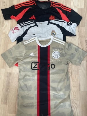Storpaket Adidas fotbollströjor - Säljer tre snygga fotbollströjor från Adidas. En beige Ajax-tröja med röda och svarta ränder och Ziggo-logga, en vit Real Madrid-tröja med klubbmärke och en svart/röd tröja med retrostil. Perfekta för träning eller match!