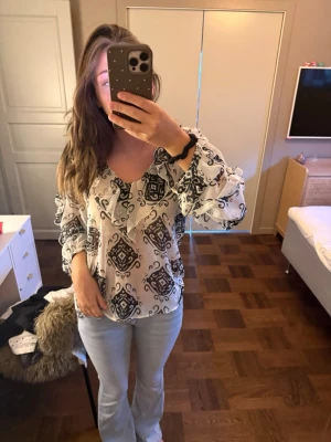 Vit blus med svart mönster och volang - Snygg vit blus med svart grafiskt mönster och volangdetaljer längs ringningen. Blusen har v-ringning och långa ärmar med lätt puff och resår vid ärmslut. Tunt, flowigt material som ger en luftig känsla. Perfekt att styla med jeans eller kjol.