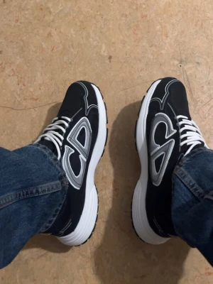 Svarta  sneakers med vita detaljer - Snygga svarta sneakers med vita och grå detaljer samt tydlig CD-logga på sidan. Skorna har vita snören och en tjock, vit sula som ger en sportig vibe. Ovandelen är i textil och syntetmaterial för en modern look.