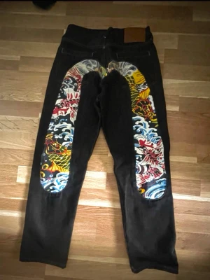 Evisu Svarta jeans  - Unika svarta jeans med färgglada japanska motiv på bakstycket. Mönstret har inslag av vågor, fiskar och drakar i rött, blått, gult och vitt. Jeansen har rak passform och klassisk femficksdesign. Perfekta för dig som vill sticka ut med en kreativ och cool stil.