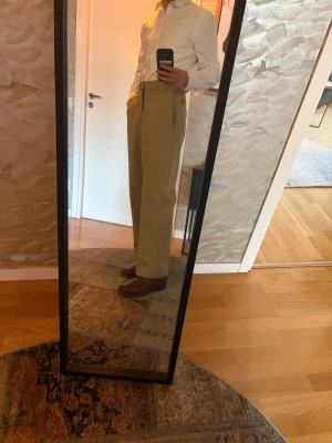 Beiga vida kostymbyxor - Snygga beiga kostymbyxor med hög midja och vida ben. Klassisk passform med pressveck framtill och diskreta sidfickor. Perfekta för en stilren look. 