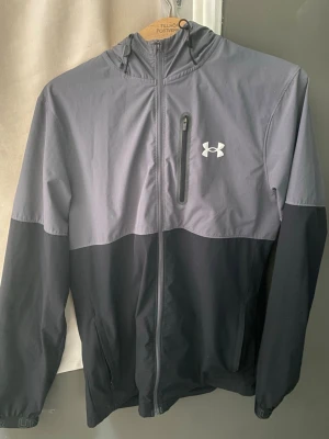 Under Armour tvåfärgad vindjacka - Snygg vindjacka från Under Armour i svart och grått. Jackan har huva, hel dragkedja framtill, två sidofickor och en bröstficka med dragkedja. Lätt material som passar perfekt för blåsiga dagar. Under Armour-logga på bröstet.