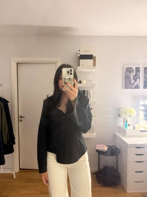 Svart omlottblus med v-ringning - Stilren svart omlottblus med v-ringning och långa ärmar. Skjortan har en figurnära passform och är tillverkad i ett mjukt, lätt tyg som ger en elegant look. Perfekt att matcha med både jeans och kostymbyxor för en trendig stil. Tröjan är för stor för mig där av säljer jag den. 