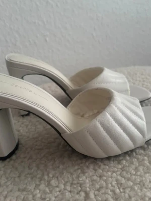 Nya. Vita quiltade sandaler från Bershka - Säljer ett par trendiga vita sandaler från Bershka med bred quiltad rem och chunky klack. Sandalerna har öppen tå och fyrkantig form, vilket ger en modern look. Materialet är syntetiskt och mjukt, perfekt för dig som gillar stilrena detaljer. Kom aldrig till användning, bara testade hemma, storlek 35, men stor i storleken och kan passa även 36, innelängd 23cm.