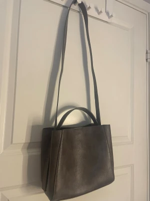 Grå brun axelväska från Gina Tricot - Grå brun axelväska från Gina Tricot. Väskan har både ett långt axelband som går att ta av och justera och ett kortare handtag, samt en innerficka med dragkedja och magnetlås. Endast använd fåtal gånger.