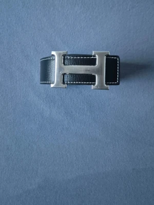 Herme's bälte 120cm - Snyggt svart läderbälte från Herme's med ikoniskt H-format metallspänne i silverfärg. Bältet har vita kontrastsömmar längs kanterna och en klassisk, stilren design som lyfter vilken outfit som helst.