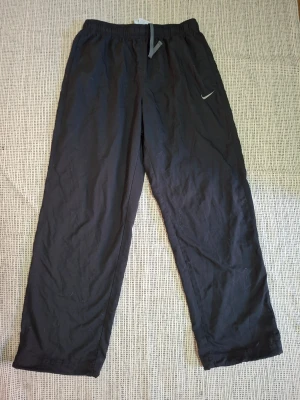 Svarta Dri-FIT byxor från Nike - Svarta sportiga mjukisbyxor från Nike med Dri-FIT-teknologi. Byxorna har resår och dragsko i midjan samt diskret Nike-logga på benet. Tillverkade i 100% polyester för en lätt och luftig känsla, perfekta för träning eller chill.