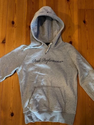 Grå hoodie från Peak Performance - Snygg grå hoodie från Peak Performance med svart broderad logga på bröstet. Klassisk känguruficka framtill, dragsko i huvan och ribbade muddar. Perfekt för en avslappnad och sportig stil. Mjuk bomullsblandning som känns skön mot huden.