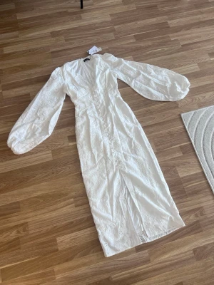 Vit långklänning med puffärmar - Superfin vit långklänning med v-ringning och broderade detaljer. Klänningen har långa puffärmar och är tillverkad i bomull, vilket gör den både luftig och bekväm. Perfekt för sommarens alla tillfällen! Dragkedja bak. Nypris 699kr!