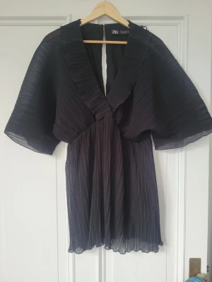 Svart plisserad kortklänning från Zara - Svart plisserad kortklänning från Zara med djup v-ringning och vida, korta ärmar. Klänningen har ett luftigt och elegant fall med markerad krage och transparenta detaljer. Perfekt nu i sommar! M men passar mig som brukar ha XS och S! Lite kort på mig som är 173, skulle säga att den fungerar för någon som är 165 ich!