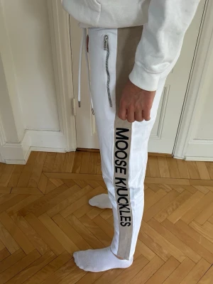 Vita mjukisbyxor Moose Knuckles - Mjukisbyxor från Moose Knuckles med beige sido mönster och svart logotyp längs benet. Byxorna har dragkedjor vid fickorna och dragsko i midjan. Slim fit-modell i mjukt och skönt material. Köpta från Zgander i Helsingborg.