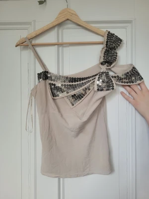 Beige paljettopp med rosett - Supersnygg beige topp med ett axelband och stor rosett på axeln. Dekorerad med glittriga silverpaljetter längs kanten och på rosetten. Delen som hålls ut kan man ha över armen🩷 från karen millen! Har några deffekter som syns i bilderna men det går nog bort med en fläckbortagare! Är i S men passar även XS och M!