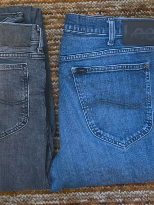 Blå och grå jeans från Lee - Två par klassiska jeans från Lee, ett i blå denim och ett i grått. Båda har raka ben, femficksmodell och tydliga sömmar. Perfekta för en avslappnad och trendig look. Materialet är bomullsdenim med snygg tvättad finish.
