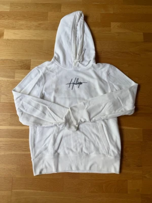 Vit hoodie från Hollister med tryck - Vit hoodie från Hollister med svart logotyp på bröstet och stort tryck med blommor och text på ryggen. Klassisk huva, känguruficka och ribbade muddar. Mjuk bomullskänsla, perfekt för kalla dagar.