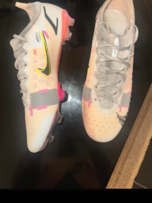 Nike Mercurial fotbollsskor beige/rosa - Säljer ett par Nike Mercurial fotbollsskor i beige med detaljer i svart, neon, rosa och grått. Skorna har snörning, låg profil och mönstrad ovandel i syntetmaterial för bra grepp på planen. Använda max 10 gånger och inte mer.