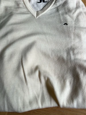 Vit stickad tröja från J.Lindeberg - Snygg vit stickad tröja från J.Lindeberg med V-ringning och diskret logga broderad på bröstet. Tröjan har ribbade muddar vid ärmslut och nederkant. Perfekt för dig som gillar stilrena och klassiska plagg.