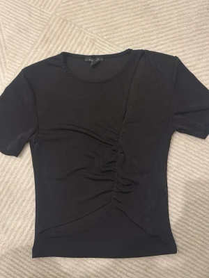 Svart topp med rynkade detaljer - Svart kortärmad topp från New yorker med rynkade detaljer framtill. Tunn och stretchig kvalitet med rund halsringning. Perfekt för dig som gillar stilrena och trendiga plagg.