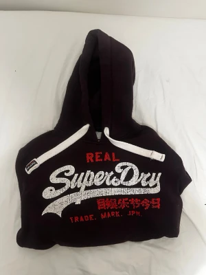 Superdry Hoodie - Snygg vinröd hoodie från Superdry med stort vitt och rött tryck på bröstet och japanska tecken. Klassisk huva med vita dragsnören och mjukt foder på insidan. Står att det är storlek XS fast passar mest storlek S.