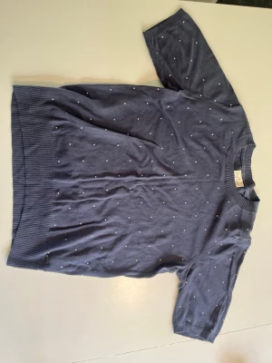 Blå stickad tröja med prickar - Snygg blå stickad tröja från Lindex med korta ärmar och små vita prickar över hela plagget. Tröjan har ribbade muddar vid hals, ärmslut och nederkant. Perfekt för dig som gillar klassisk och stilren design.