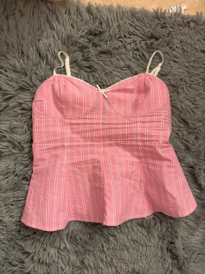Rosa randig korsett-topp med rosett - Supersöt rosa korsett-topp med vita ränder och tunna axelband. Toppen har en liten rosett framtill och markerade kupor för en feminin look. Perfekt att styla med jeans eller kjol för en trendig outfit.