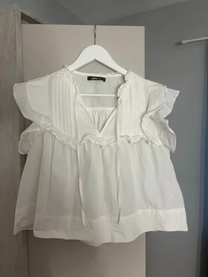 Vit blus med volang/bluzka - Söt vit blus från Gina Tricot med volangdetaljer och knytband vid halsen. Blusen har korta ärmar med rynk och dekorativa sömmar framtill. Perfekt för en trendig och avslappnad look.