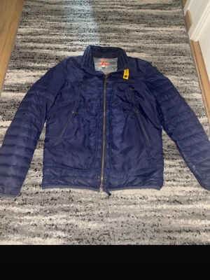 Blå vindjacka från Parajumpers - Snygg blå vindjacka från Parajumpers med klassisk gul detalj vid kragen. Jackan har dragkedja framtill, två snedställda fickor med dragkedja och quiltade ärmar. Materialet är glansigt och lätt, perfekt för kalla dagar.