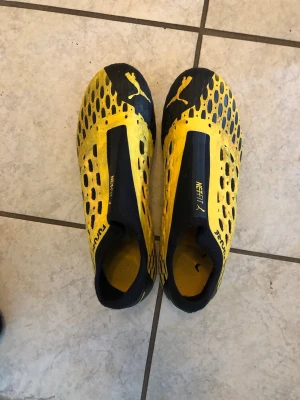 Puma gul-svarta fotbollsskor - Säljer ett par grymma Puma Netfit fotbollsskor i svart och gult. Skorna har en modern design med mönstrad ovandel och elastisk snörning för optimal passform. Perfekta för dig som vill sticka ut på planen och få bra grepp på gräs.