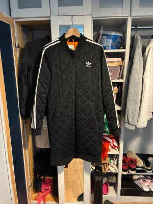 Svart quiltad lång jacka från Adidas - Svart lång quiltad jacka från Adidas med vita ränder längs ärmarna och dragkedja framtill. Jackan har ribbade muddar och klassisk Adidas-logga på bröstet. Perfekt för kyliga dagar och har en sportig streetstyle vibe.