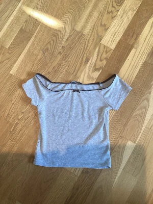 Grå offshoulder topp - Snygg grå offshoulder topp med svarta spetsdetaljer längs kanten och en liten rosett framtill. Toppen är kortärmad och har en tight passform, perfekt för en trendig look. Storlek S men kan absolut andvändas som xxs!