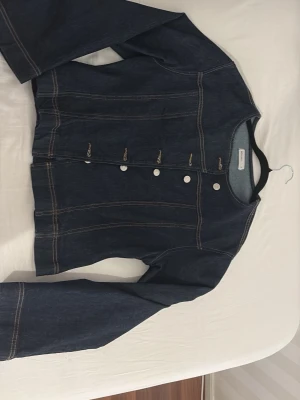 Mörkblå jeansjacka från Gina Tricot - Snygg mörkblå jeansjacka från Gina Tricot med raka sömmar och silverfärgade knappar framtill. Jackan har en klassisk, rak passform och kontrastsömmar i gult som ger en cool vintagekänsla. Perfekt för dig som vill ha en stilren och tidlös jeansjacka.