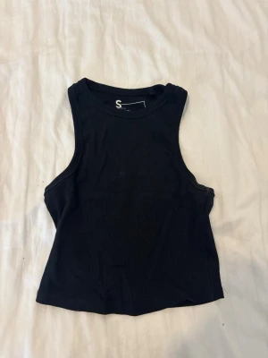 Svart ribbad linne - En svart, croppad linnetopp i ribbad bomull med ärmlös design och rund halsringning. Perfekt för att styla med jeans eller kjol för en trendig look.