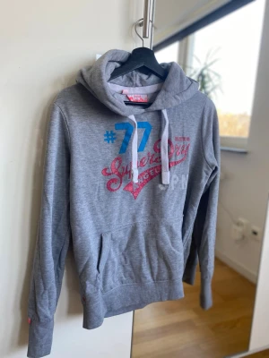 Grå Superdry hoodie med tryck - Snygg grå hoodie från Superdry med stort tryck i blått, rött och vitt framtill. Klassisk huva med vita snören och mjukt material. Perfekt för en avslappnad streetstyle-look.