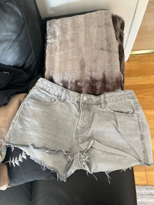 Grå denimshorts med råa kanter - Säljer ett par grå denimshorts med råa, fransiga kanter och klassisk femficksdesign. Shortsen har hög midja och stängs med knappar  Perfekta för sommardagar och ger en avslappnad look.