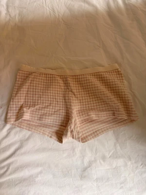 Rutiga ljusrosa shorts med resår - Säljer ett par ljusrosa shorts med rutigt mönster och bred resår i midjan. De är tillverkade i ett mjukt och lätt tyg, perfekta för varma dagar eller som mysplagg hemma. Passar dig som gillar en avslappnad och trendig stil.