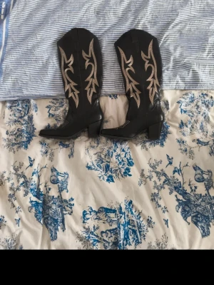 Svarta cowboy boots med broderi - Snygga svarta cowboy boots med beige broderade detaljer och klassisk western-design. Bootsens ovandel är i skinn och de har en stadig klack. Perfekta för dig som vill ha en cool westernstil med extra detaljer.Vet ej om de är äkta skinn,Inget kvitto 