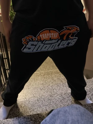 Trapstar Shooters svarta mjukisbyxor - Svarta mjukisbyxor från Trapstar med stort tryck framtill: basketboll, tiger och texten 'London Shooters'. Byxorna har avslappnad passform och resår i midjan. Perfekta för chill eller streetwear-stil.