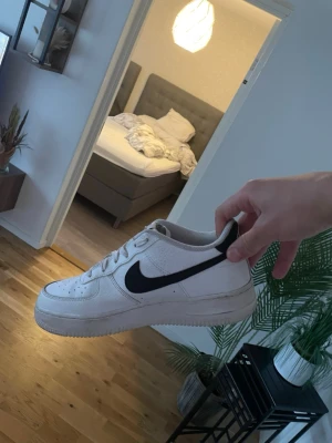 Nike Air Force 1 vita sneakers - Klassiska vita Nike Air Force 1 sneakers med svart swoosh på sidan. Skorna har lågt skaft, perforerad tåbox och vita skosnören. Tillverkade i läder med gummisula och ikonisk design som passar till det mesta.