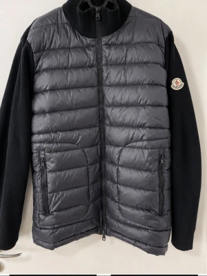 Svart cardiganjacka från Moncler - Snygg svart cardiganjacka från Moncler med quiltad framsida i dun och stickade ärmar samt rygg. Dragkedja framtill, två fickor med dragkedja och Moncler-logga på ärmen. Perfekt för lager-på-lager och streetstyle. Använd en gång och säljs pågrund av att den inte passar längre. ⚠️Pris kan disskuteras vid snabbköp, OBS byten kan accepteras i vissa fall⚠️