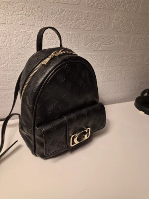Guess svart ryggsäck - Svart ryggsäck med justerbara axelband. Väskan har ett fack med dragkedja och ett mindre fack framtill som stängs med tryckknapp. Väskan har ett mönster med logotyper. 27x25x10cm