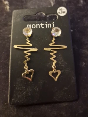 Guldiga örhängen med hjärta Montini - Snygga guldiga örhängen med glittrande stenar upptill, vågig design och ett hängande hjärta längst ner. Perfekta för att ge din look en unik touch. Materialet är metall med en klar sten som detalj.