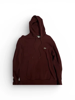 Vinröd hoodie från Lacoste - Snygg vinröd hoodie från Lacoste med klassisk krokodillogga på bröstet. Hoodien har huva med dragsko och en stor magficka framtill. Tillverkad i mjukt bomullsmaterial som är skönt att ha på sig.