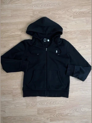Svart hoodie från Polo Ralph Lauren - Svart hoodie med dragkedja från Polo Ralph Lauren. Klassisk design med huva och dragsnören, två fickor framtill och den ikoniska vita logon broderad på bröstet. Tillverkad i mjukt bomullsmaterial som är skönt att bära.