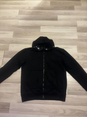 Svart hoodie från C.P. Company - Säljer en svart hoodie från C.P. Company med dragkedja och ikoniska glasögon i huvan. Hoodien har broderad logga på bröstet och stora fickor framtill. Tillverkad i mjukt bomullsmaterial och har en clean streetwear-look.