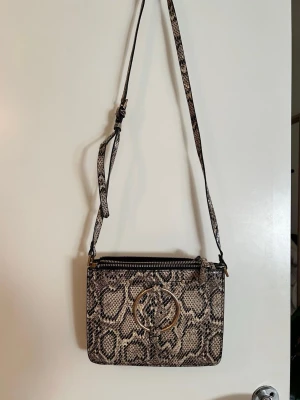 Axelväska i ormmönstrat konstläder - Snygg axelväska i beige och svart ormmönstrat konstläder med justerbar rem och guldfärgad ringdetalj framtill. Väskan har två fack med dragkedja och svart innerfoder. Perfekt för dig som vill ha en trendig accessoar med plats för det viktigaste.