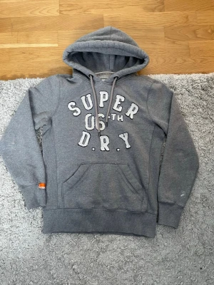 Grå hoodie från Superdry - Snygg grå hoodie från Superdry, bra skick förutom på armbågarna där de har fått någon slags av färg, men det är inget man tänker på när man har på sig tröjan, annars i jättebra skick, Syns inte jättetydligt! Det är i st S, priset är inte spikat, MVH ReStyle.Fashion🤗