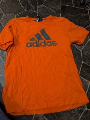 Orange Adidas t-shirt med logga stl 13-14 år - Snygg orange t-shirt från Adidas med stor svart logga på bröstet. Klassisk rund hals och korta ärmar. Tillverkad i mjuk bomull som känns skön mot huden. Perfekt för dig som gillar sportig och avslappnad stil.