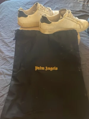 Palm Angels vita sneakers med svarta detaljer - Palm Angels sneakers i vitt skinn med svarta detaljer på hälen och klassisk snörning. Märkets logga syns på sidan och skorna har en stilren, låg profil. Perfekta för dig som gillar streetwear och vill ha en clean look. Pris kan diskuteras 
